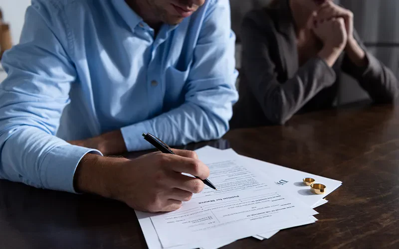 A man in a blue button-down shirt signing divorce papers at a wooden table while a woman in a grey blazer sits in the background with her hands clasped, next to two gold wedding rings.