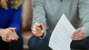 A close-up, shallow-depth-of-field shot of a person holding a printed document and pointing a pen toward it while sitting next to another individual.