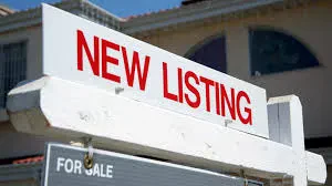 A white real estate signpost features a prominent top rider reading "NEW LISTING" in bold red capital letters, with a gray "FOR SALE" sign hanging underneath.
