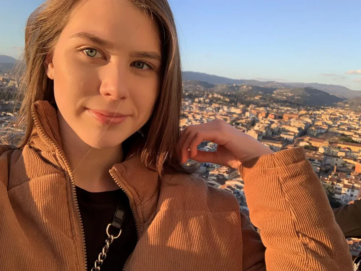 A close-up selfie of Sydney. A woman with light green eyes wearing a brown corduroy jacket, with a sprawling European city layout nestled in hills behind her at sunset