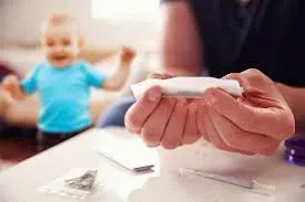 Close-up of a person's hands rolling a marijuana joint over a white table, with a blurred toddler in a blue shirt playing in the background.