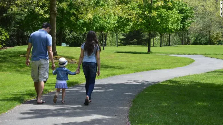 Rear view of a family of three—a man, a woman, and a young child wearing a white hat—holding hands while walking down a paved, winding path in a sunny, green park.