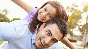 A man and a young girl smile together while sitting on the grass, enjoying a sunny day outdoors.