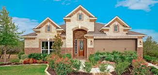 A single-story house with tan brick and stone exterior, a brown shingled roof, a landscaped front yard with shrubs and flowers, two-car garage, and blue sky with scattered clouds in the background.