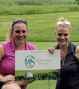 Attorneys Kristen Wolf and Shari Shore smiling on a grassy golf course while holding a white sponsorship sign that features the green Wolf and Shore Law Group logo