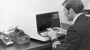 A man in a suit sits at a desk, focused on his computer, with a professional office environment in the background.