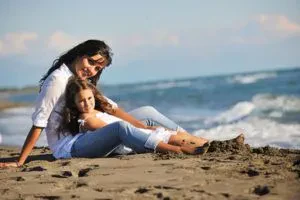 A woman and a young girl sit together on a sandy beach, smiling at the camera. The ocean waves are in the background, and both are wearing white tops and jeans.