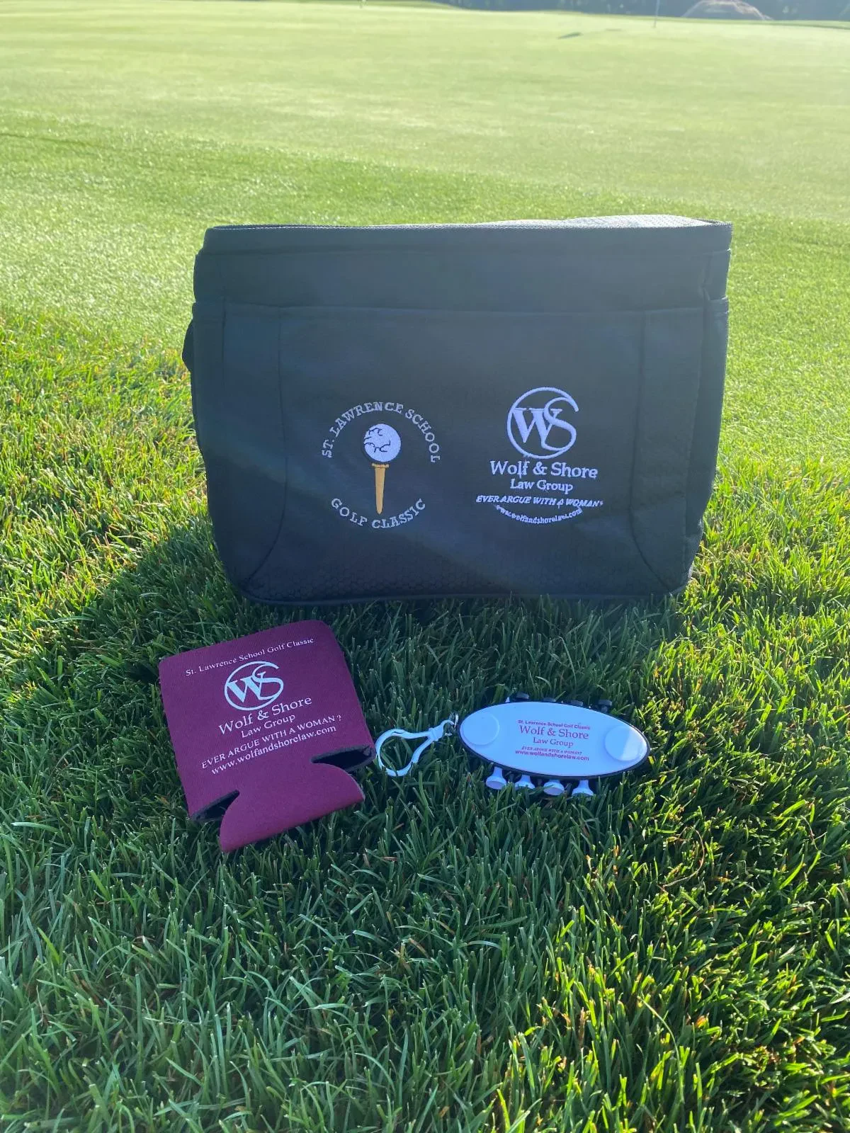 Custom branded golf swag layout on green grass, featuring a black cooler bag, a maroon can koozie, and a white golf tee holder combo printed with the St. Lawrence School Golf Classic and Wolf & Shore Law Group logos.