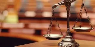 A bronze balance scale of justice positioned on a wooden desk with a blurry background of a classic courtroom setting.