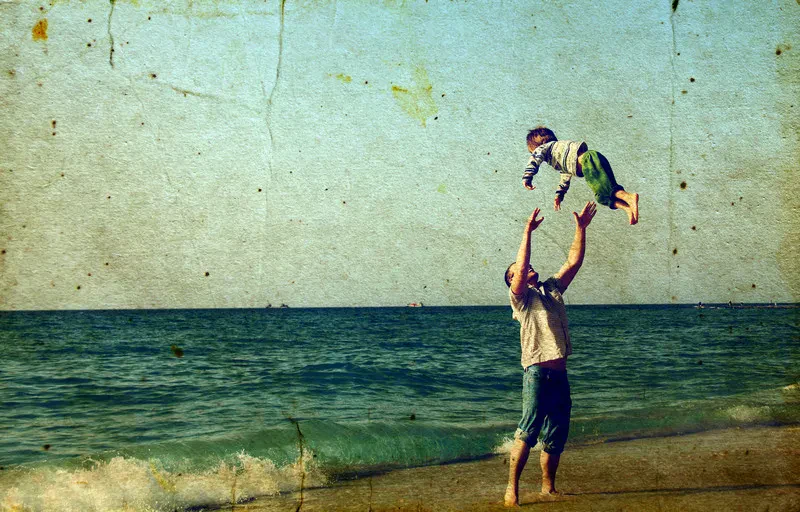 A father throws his young child into the air on a sandy beach by the ocean, styled with a textured vintage filter.