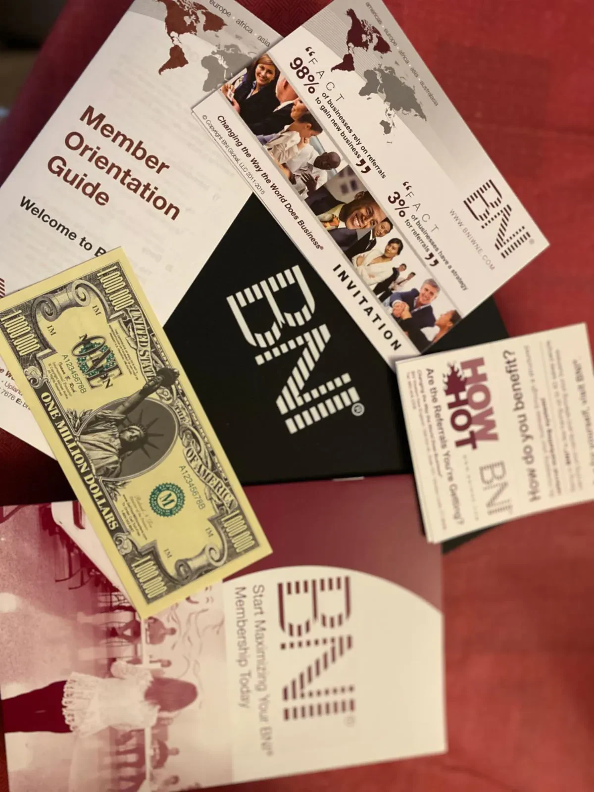 BNI marketing materials and a Member Orientation Guide arranged on a red surface, featuring an invitation card, informational brochures, and a novelty million-dollar bill.