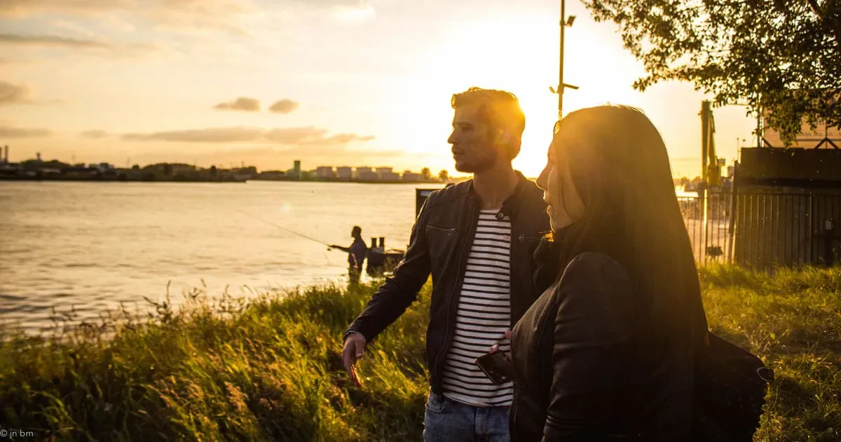 A man in a striped shirt and leather jacket stands next to a woman along a grassy riverbank, both looking out toward the water during a golden hour sunset.