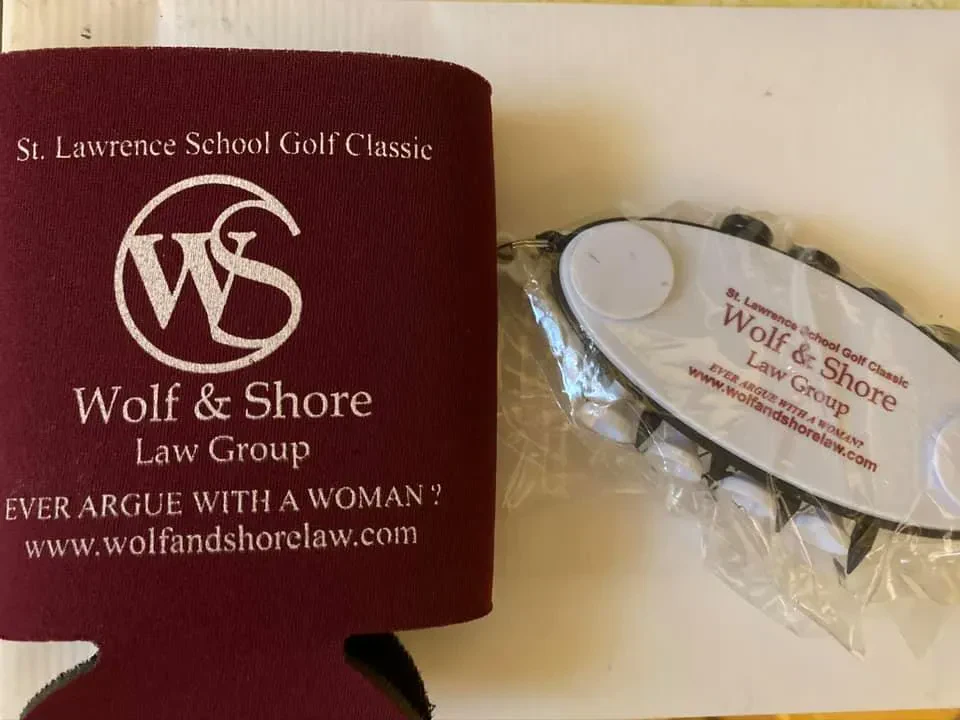 A maroon can koozie and a packaged white golf accessory item printed with the St. Lawrence School Golf Classic and Wolf and Shore Law Group logos alongside the text EVER ARGUE WITH A WOMAN?