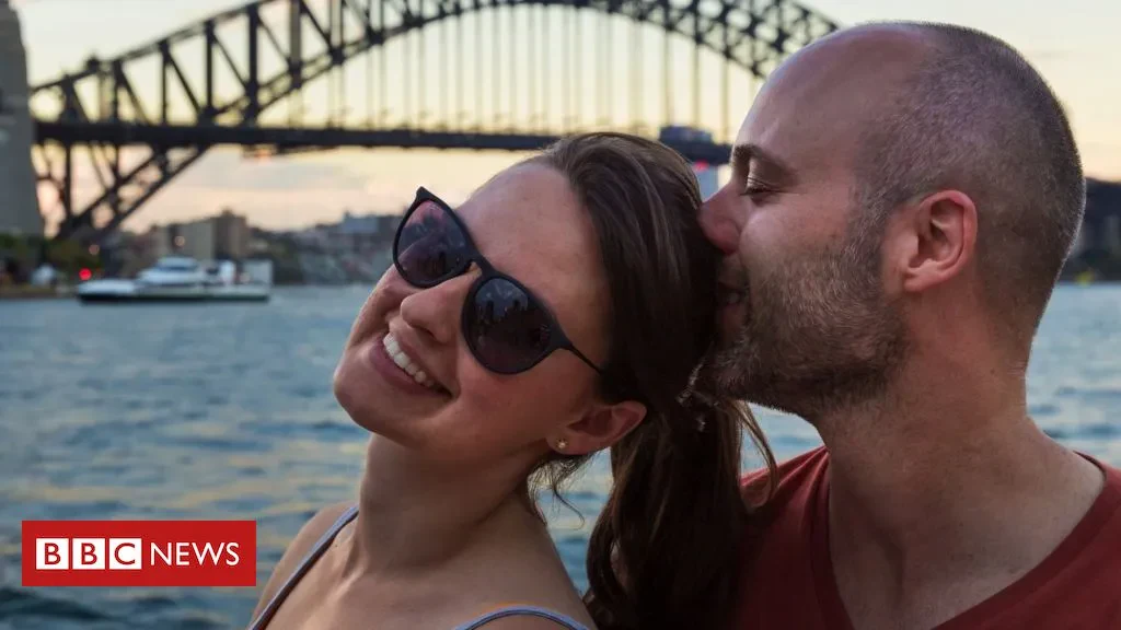 A man kissing a smiling woman wearing sunglasses on the head, with the Sydney Harbour Bridge and water in the background, featuring a BBC News logo overlay.
