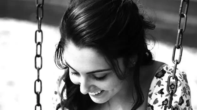 A close-up black and white photograph of a smiling young woman looking down, framed on both sides by the metal chains of a swing set.