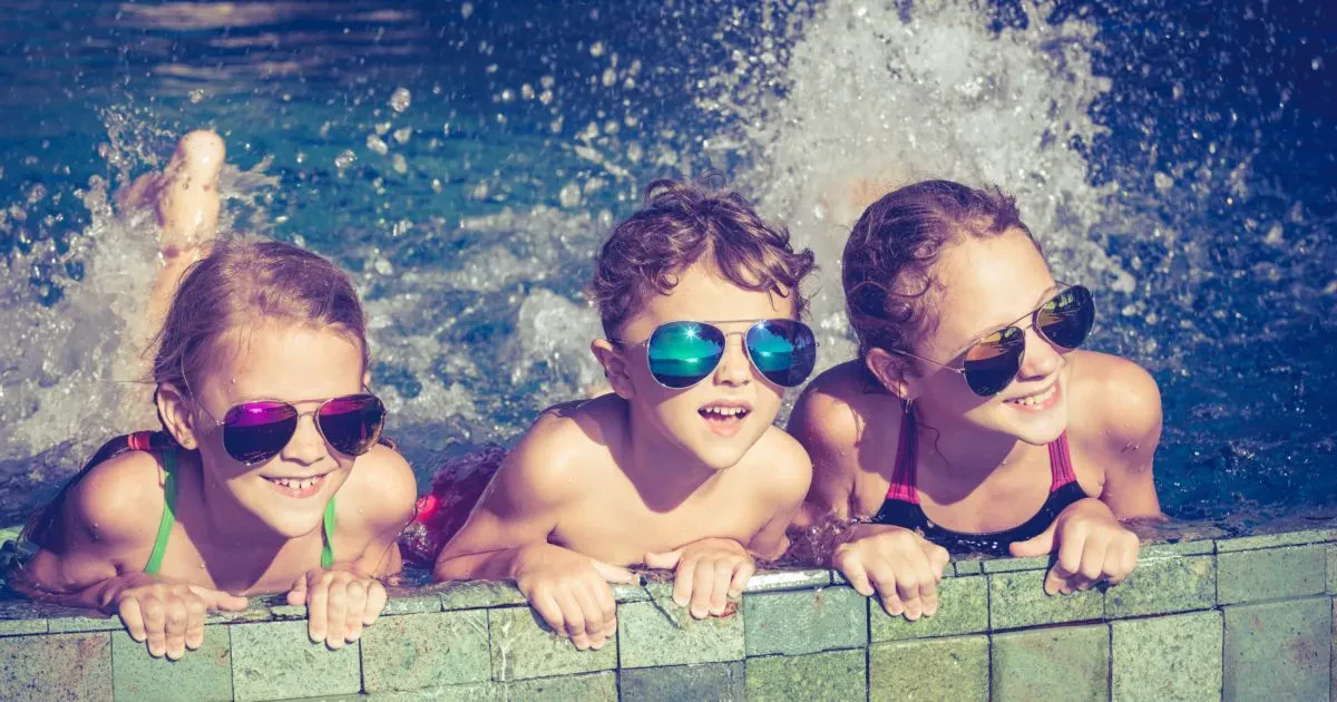 Three young children wearing sunglasses lean over the tiled edge of a swimming pool, smiling and splashing in the water.