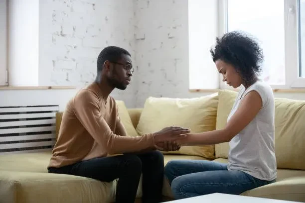 An African American man with glasses and a tan sweater sits on a yellow sofa, gently holding the hands of an African American woman in a white t-shirt who is looking down with a sad or pensive expression.