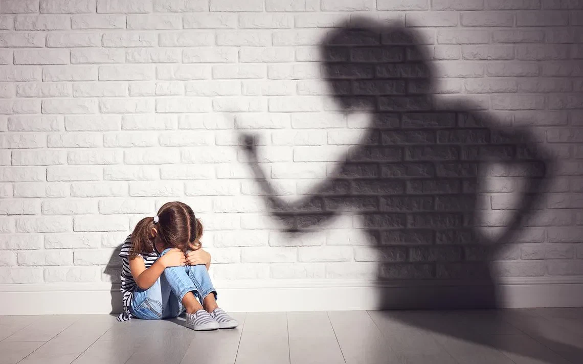 A young girl sits on the floor with hands on her knees, while her mother's shadow is cast on the wall behind her.