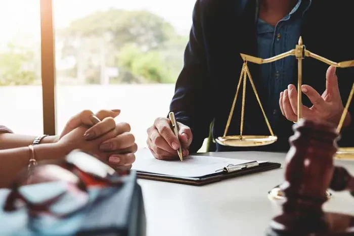 A lawyer or legal professional holding a pen over documents on a clipboard while discussing a case with a client who has their hands clasped across the desk, with a golden scale of justice and a wooden judge's gavel visible in the foreground.