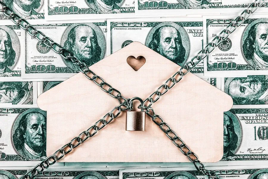 A wooden house cutout chained and padlocked over a background of one-hundred-dollar bills.