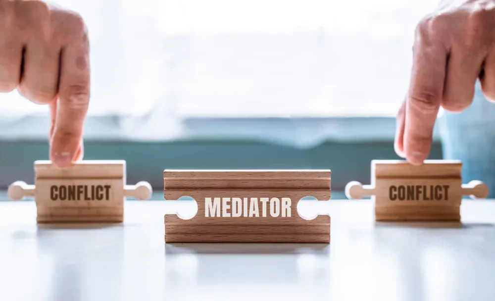 Two hands each point at wooden blocks labeled “CONFLICT,” with a central puzzle piece between them labeled “MEDIATOR,” symbolizing mediation between conflicts.