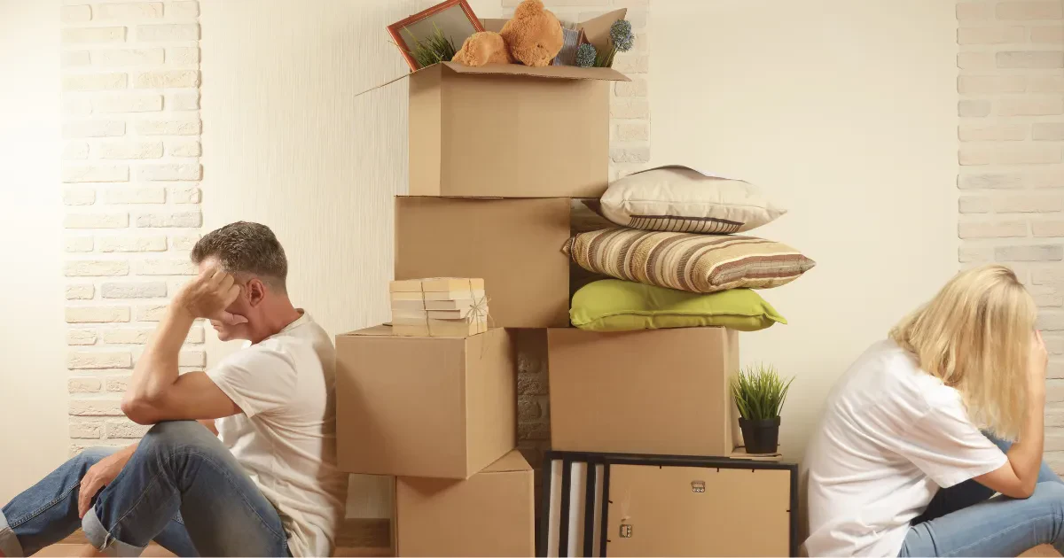 A man and woman sit back-to-back looking upset, surrounded by stacked moving boxes, pillows, and household items against a wall.