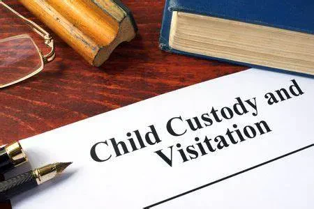 An legal document titled "Child Custody and Visitation" lying on a wooden desk next to a book, eyeglasses, and a fountain pen.