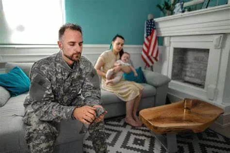 A soldier in camouflage uniform sits on a sofa, looking down pensively at his hands, while a woman sits in the background holding a baby, next to an American flag and a fireplace.