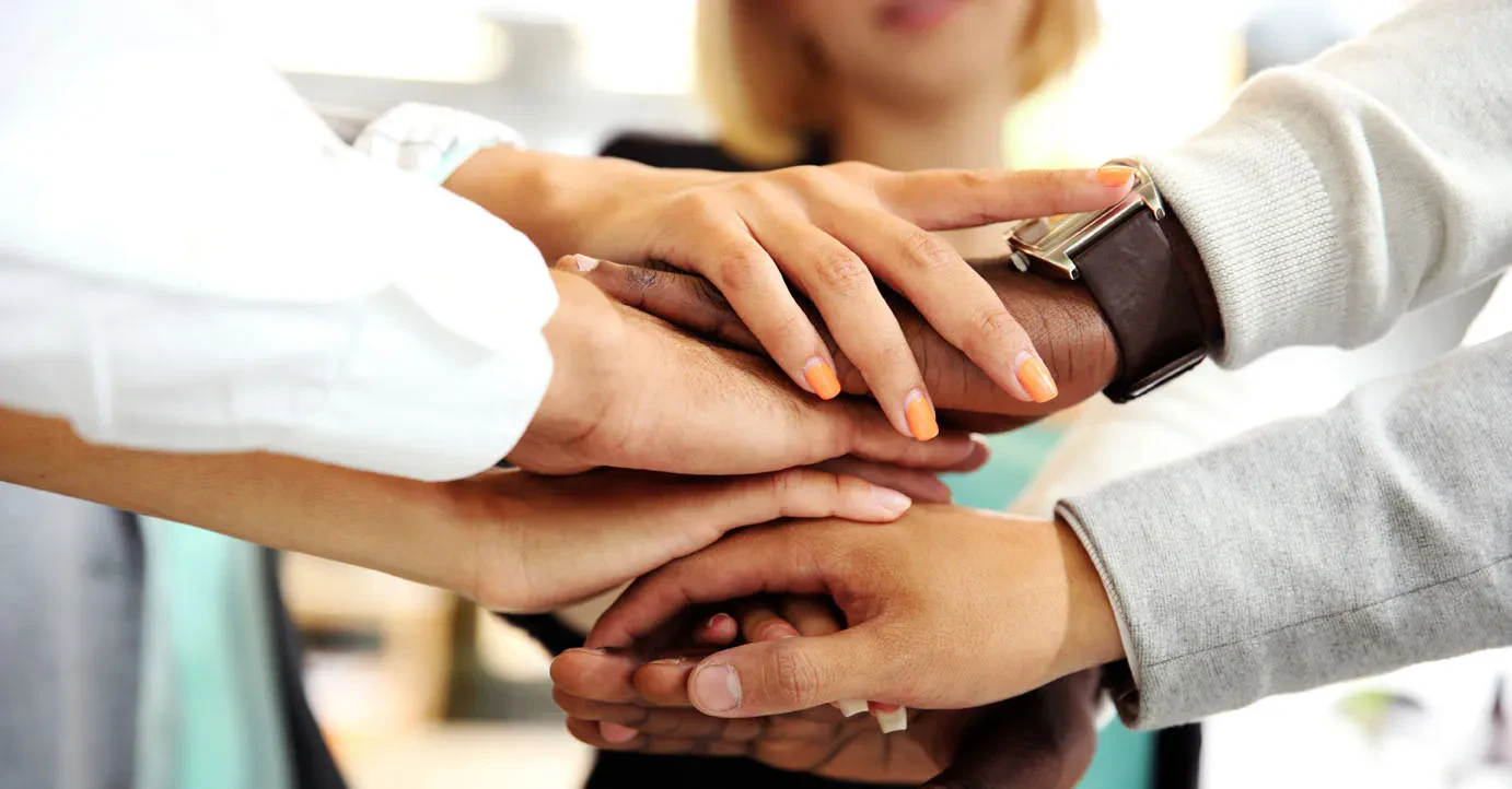 A group of people stack their hands together in a gesture of teamwork and unity, with diverse skin tones visible, symbolizing cooperation and collaboration.