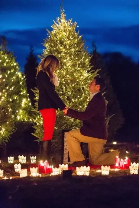 A man kneels to propose to his girlfriend in front of a beautifully decorated Christmas tree, capturing a romantic moment.