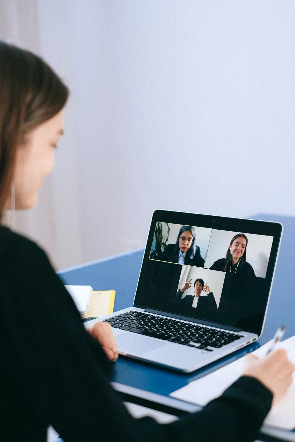 An over-the-shoulder view of a woman on a laptop hosting a three-panel video conference call with other business professionals while writing in a notebook.