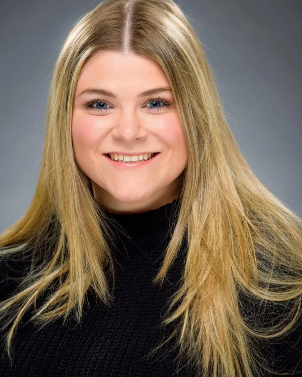 A professional studio headshot of a smiling young woman with long blonde hair and blue eyes, wearing a black knit sweater against a gradient grey background