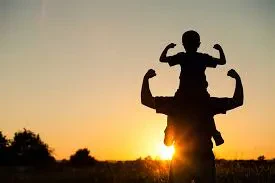 Silhouette of a child sitting on a man's shoulders, with both flexing their biceps against a bright sunset background.