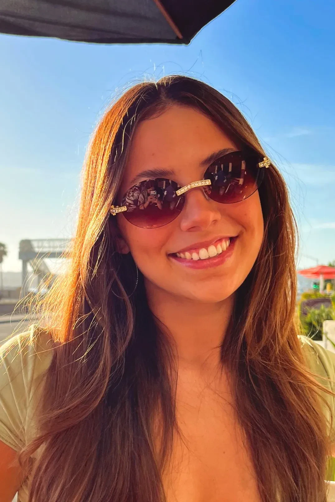 A close-up portrait of Janessa Pompano. A woman with long brown hair, wearing rimless tinted sunglasses with gold accents under a bright blue sky.