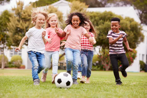 A diverse group of five happy young children running and playing with a soccer ball on a grassy field outside.