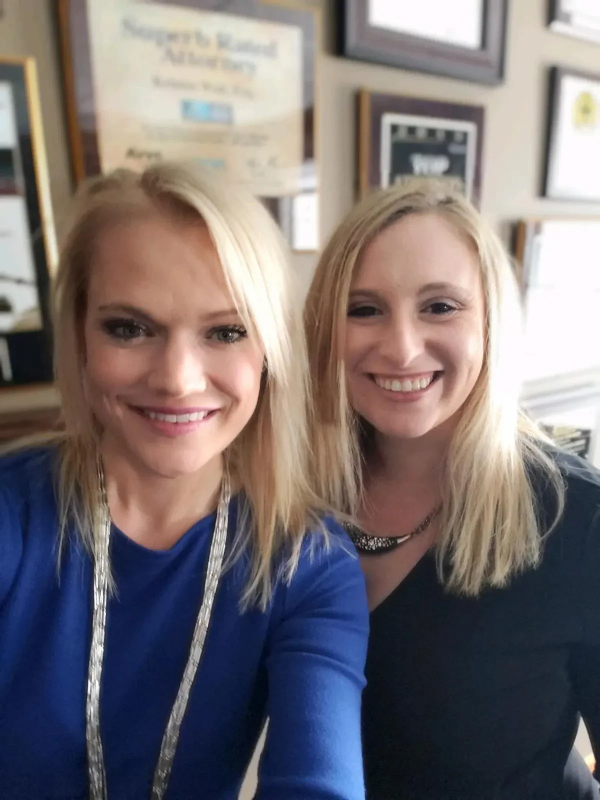 Attorneys Kristen Wolf and Shari Shore smiling side-by-side inside an office with framed legal credentials and a Superb Rated Attorney certificate hanging on the wall behind them