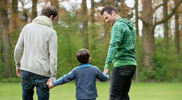 Two fathers holding hands with their young son while walking together in a grassy park surrounded by trees.