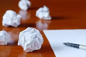 A blank sheet of white paper and a pen on a wooden desk, surrounded by several crumpled balls of paper scattered in the background.