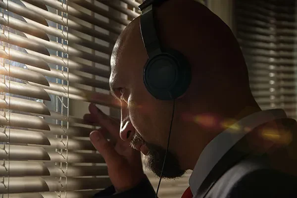 A man peeking through window blinds while wearing headphones.
