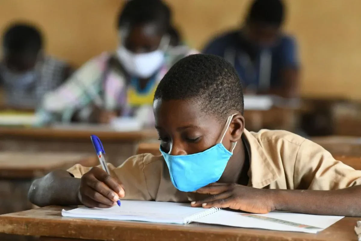 A close-up of a young boy wearing a bright blue cloth face mask while carefully writing in a notebook with a blue pen at a school desk, with other masked students blurred in the background