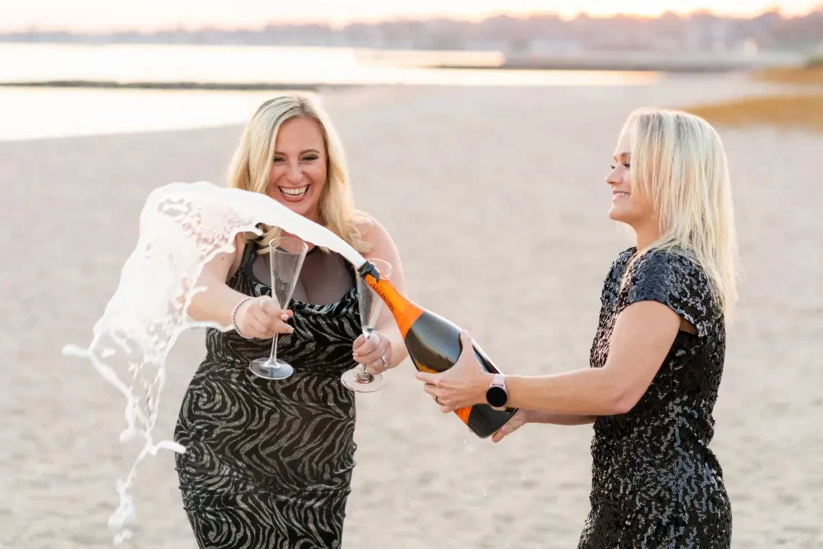 Kristen Wolf and Shari Shore in sparkly dresses celebrate on a beach at dusk while popping an orange-labeled bottle of Veuve Clicquot champagne.