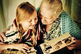 A smiling young girl with pigtails and her elderly grandmother looking through an old family photo album together.