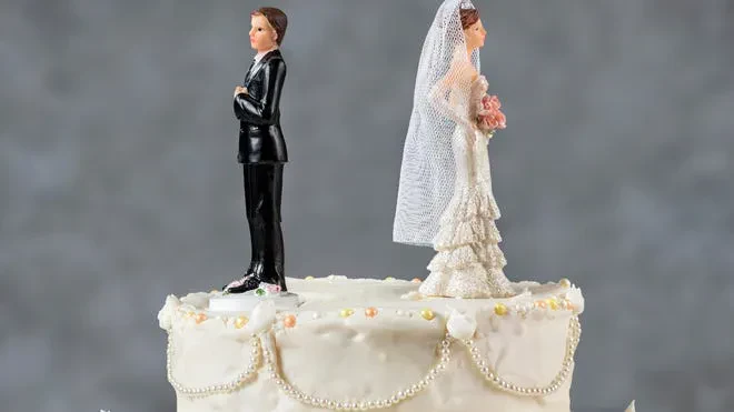 The image depicts a white, decorated wedding cake with a traditional bride and groom topper, but the two figures are turned completely away from each other with their backs facing one another.