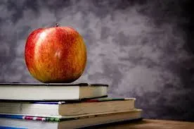 A red and yellow apple sits on top of a stack of three books, with a gray textured background.