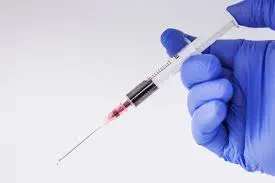 A gloved hand holds a syringe filled with blood, the needle pointing downward against a plain background.