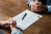 Two people sit across a wooden table from each other with their hands clasped, facing a legal document with a pen and a wedding ring resting on it.