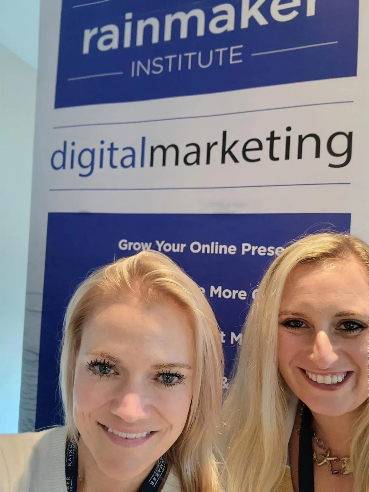 Kristen Wolf and Shari Shore take a close-up selfie in front of a blue and white banner that reads 'rainmaker INSTITUTE digitalmarketing'.