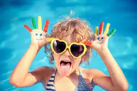 A young child in a pool wearing yellow heart-shaped sunglasses, sticking their tongue out, and holding up hands painted with bright colors and smiley faces.