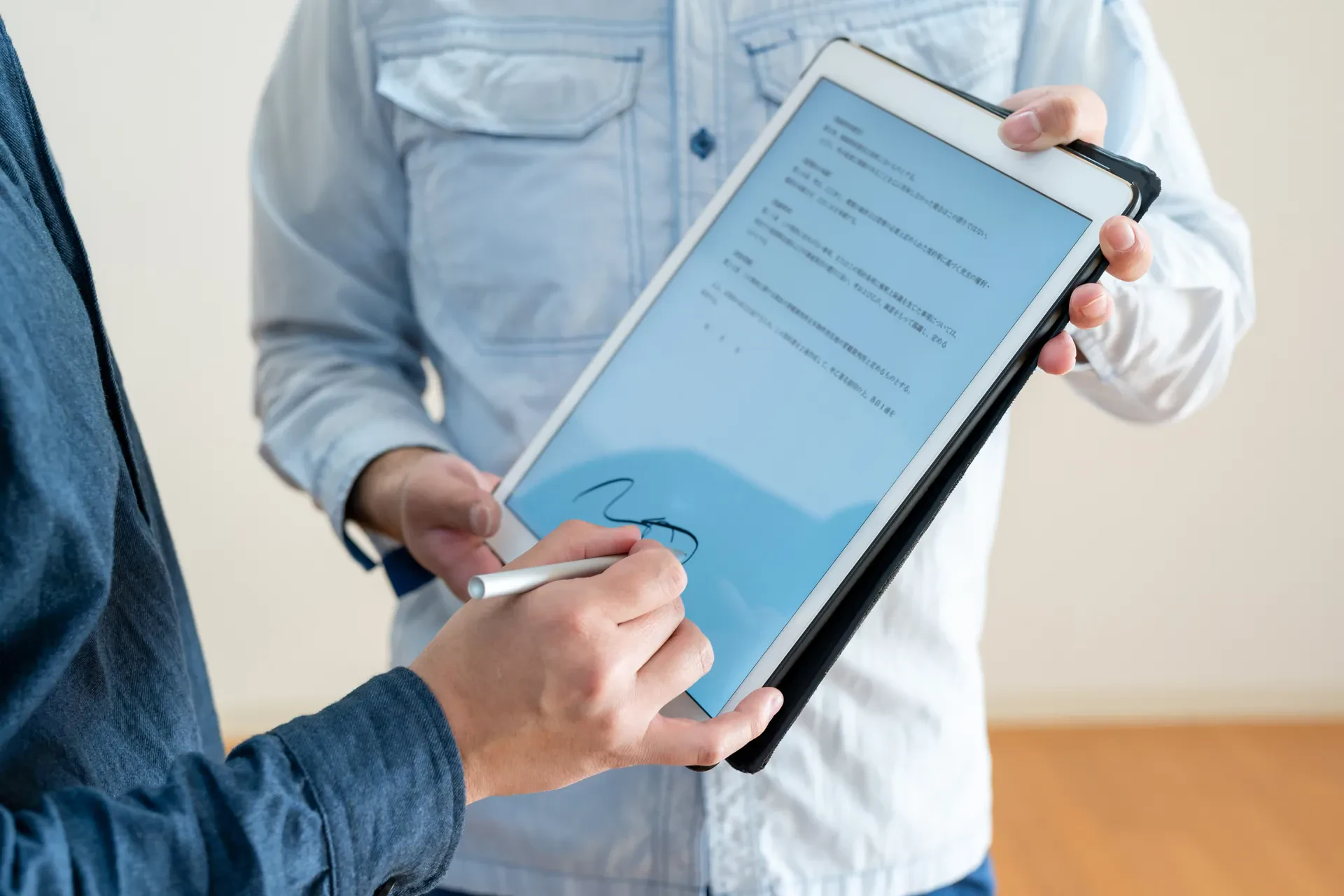An adult signs a digital document on a tablet with a stylus, while another person holds the device.