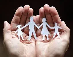 Two open hands gently hold paper cutouts of a family of four, including two adults and two children, against a dark background.